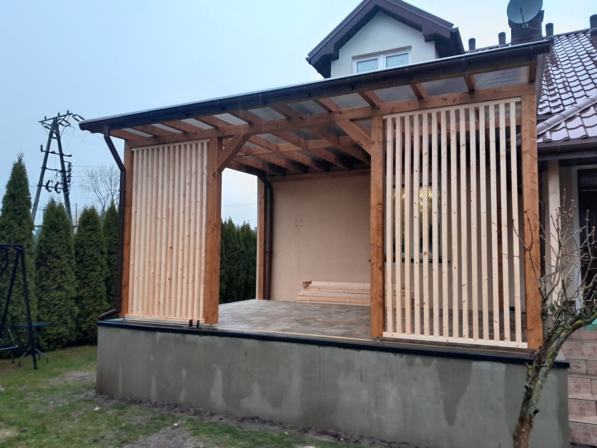 Nowoczesna drewniana pergola z lamelami, zadaszona poliwęglanem, przylegająca do domu. Widoczne elementy konstrukcyjne i surowe drewno. Częściowo ukończony taras.