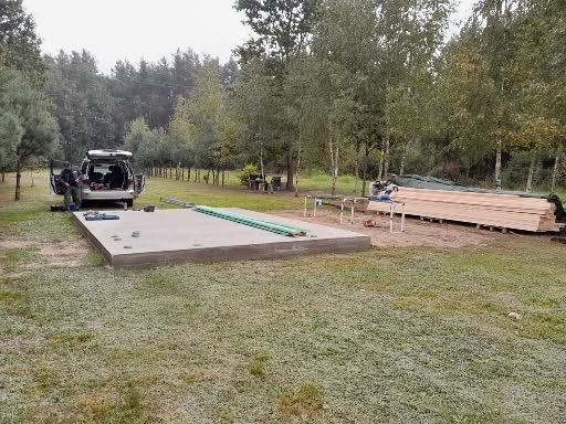 Przygotowanie do budowy tarasu drewnianego: betonowa podstawa, deski, piła stołowa, samochód dostawczy i osoba w tle, na tle drzew.