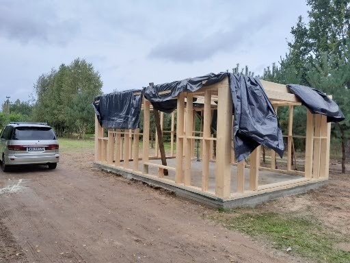 Surowa konstrukcja drewniana z czarną folią ochronną, widok z zewnątrz, obok zaparkowany samochód osobowy, teren budowy.