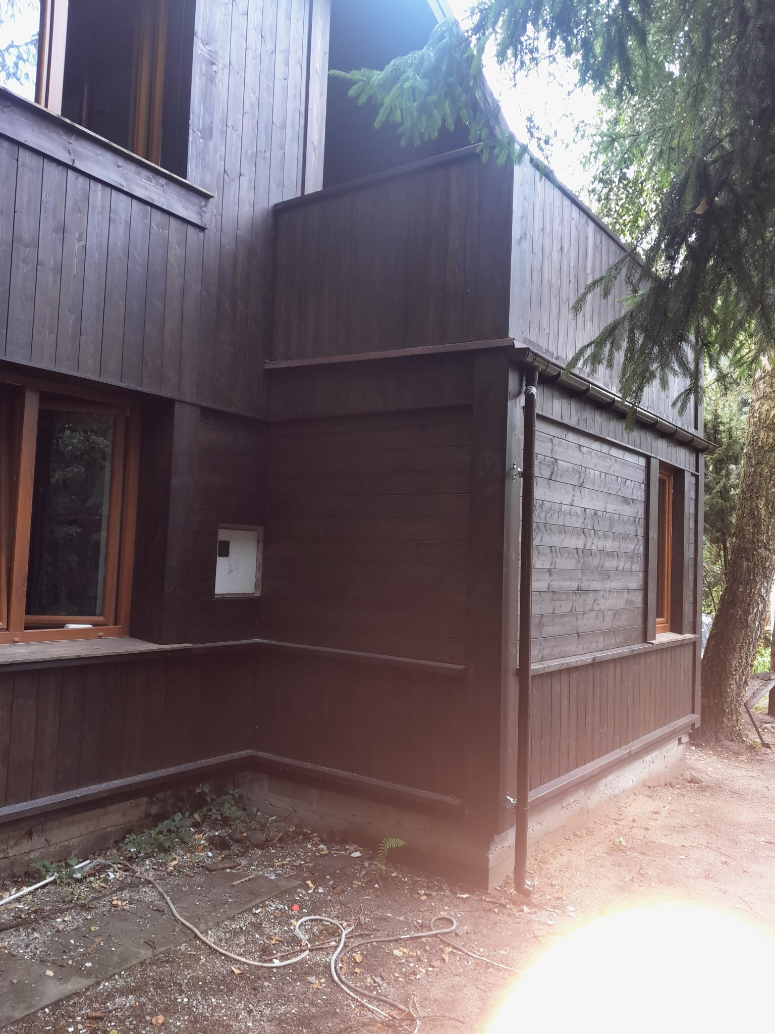 Dwukondygnacyjny budynek z ciemnego drewna z oknami w brązowych ramach, otoczony zielenią. Widok z boku, z widocznym fragmentem balkonu i rynną.