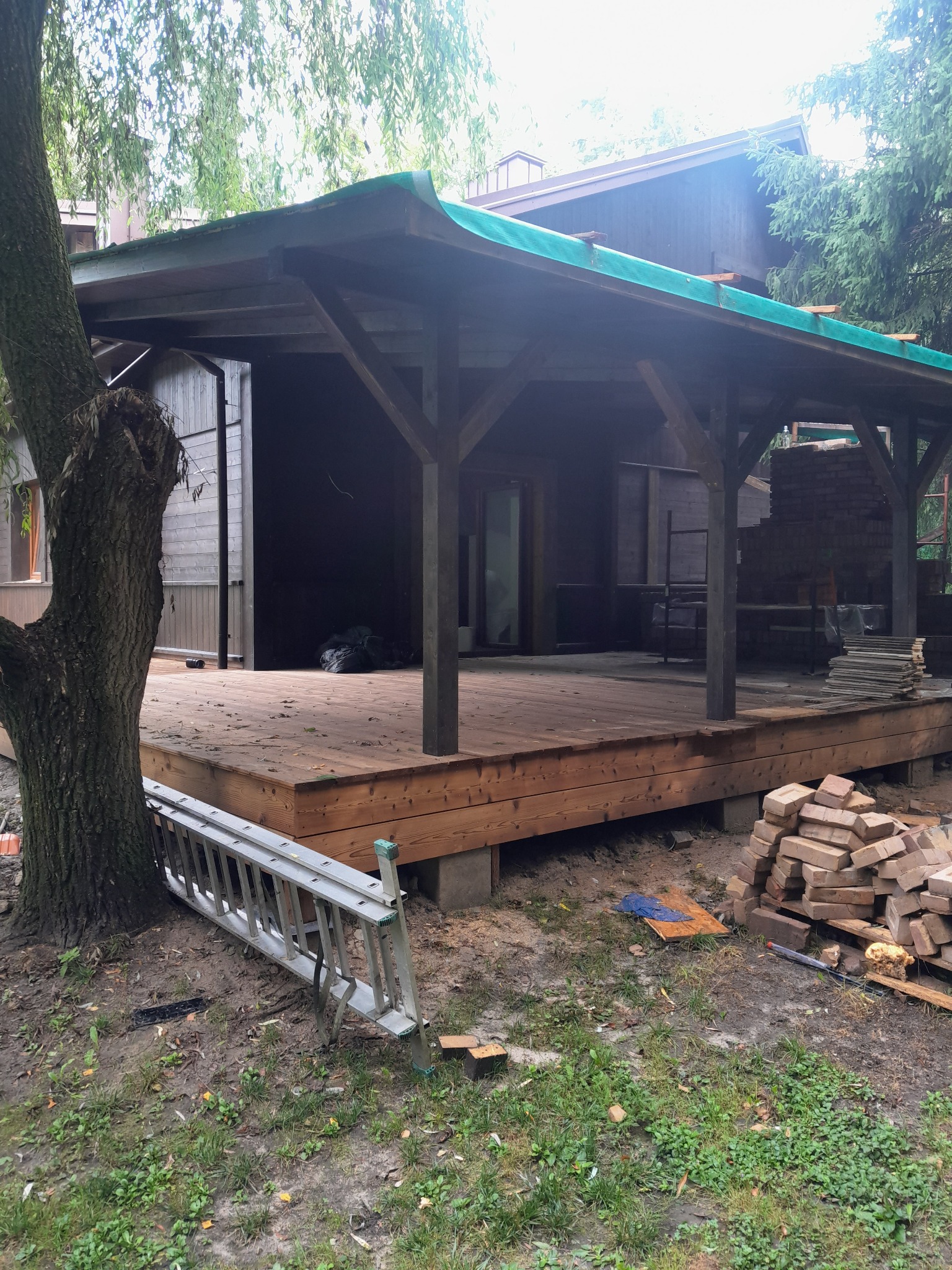 Drewniany taras z zadaszeniem w trakcie budowy, widoczne materiały budowlane i drabina. Ujęcie z poziomu gruntu, obok drzewo.