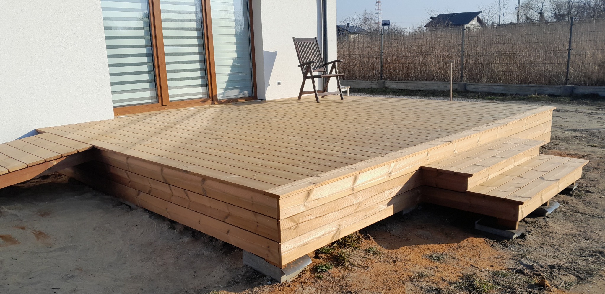 Jasny, drewniany taras z niskimi schodami i rampą przy białym budynku. Na tarasie stoi krzesło. W tle widoczne ogrodzenie i zabudowa.