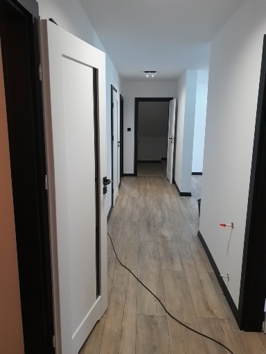 Wykończony korytarz z panelami podłogowymi w odcieniach szarości i białymi drzwiami z czarnymi ościeżnicami. Widoczne detale wykończeniowe i oświetlenie sufitowe.