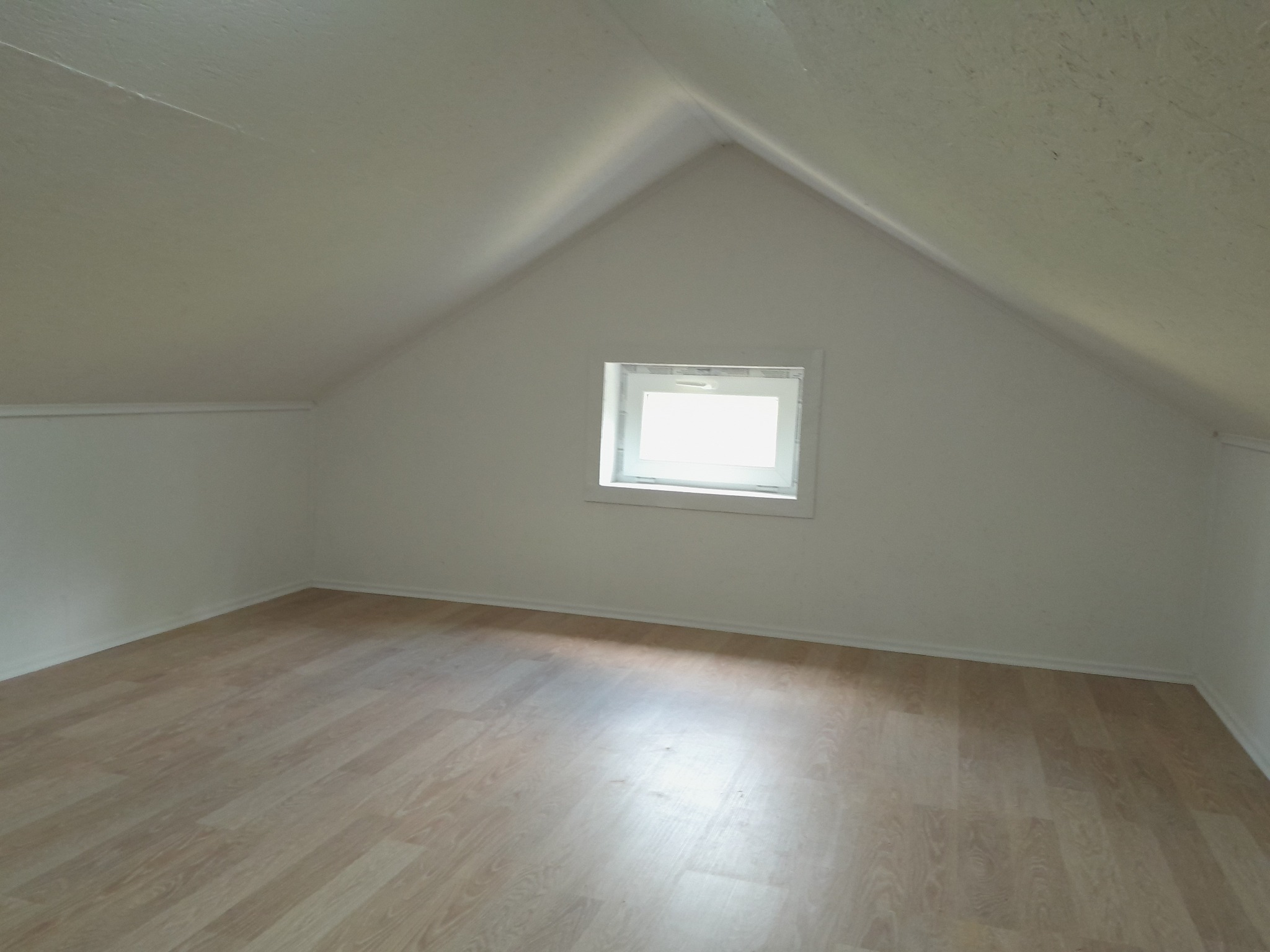 Poddasze z oknem po remoncie: jasne ściany, panele podłogowe w odcieniach beżu, skosy. Widok na puste, odnowione wnętrze.
