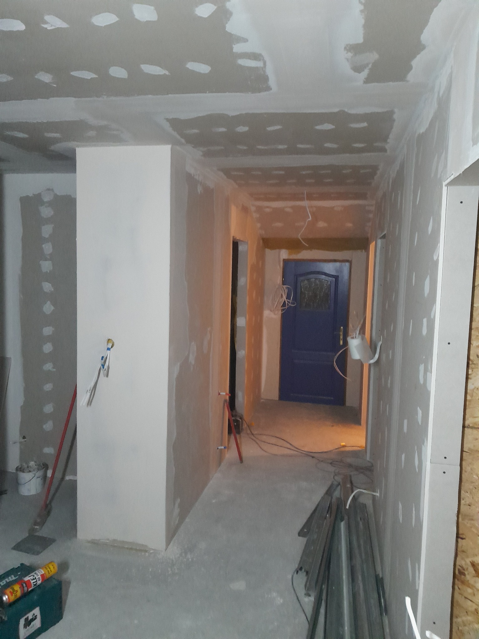 Wnętrze w trakcie prac wykończeniowych: ściany z płyt gipsowo-kartonowych, widoczne łączenia do szpachlowania, niebieskie drzwi w głębi korytarza, narzędzia na podłodze.