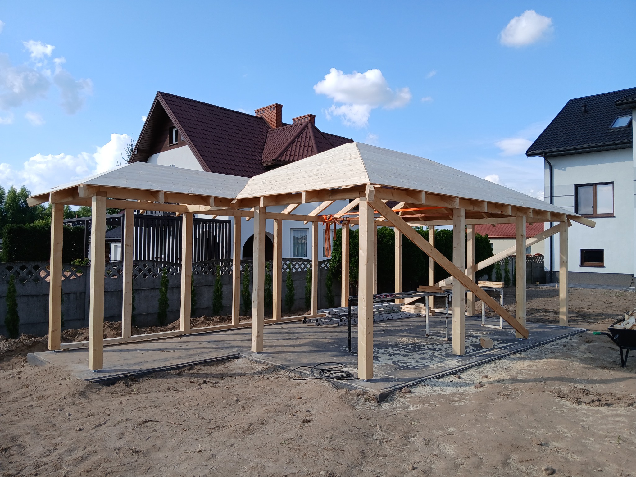 Wznoszenie drewnianej konstrukcji tarasu na betonowej wylewce. Widoczne słupy, zadaszenie w trakcie budowy, narzędzia i otoczenie posesji. Jasne drewno, błękitne niebo.