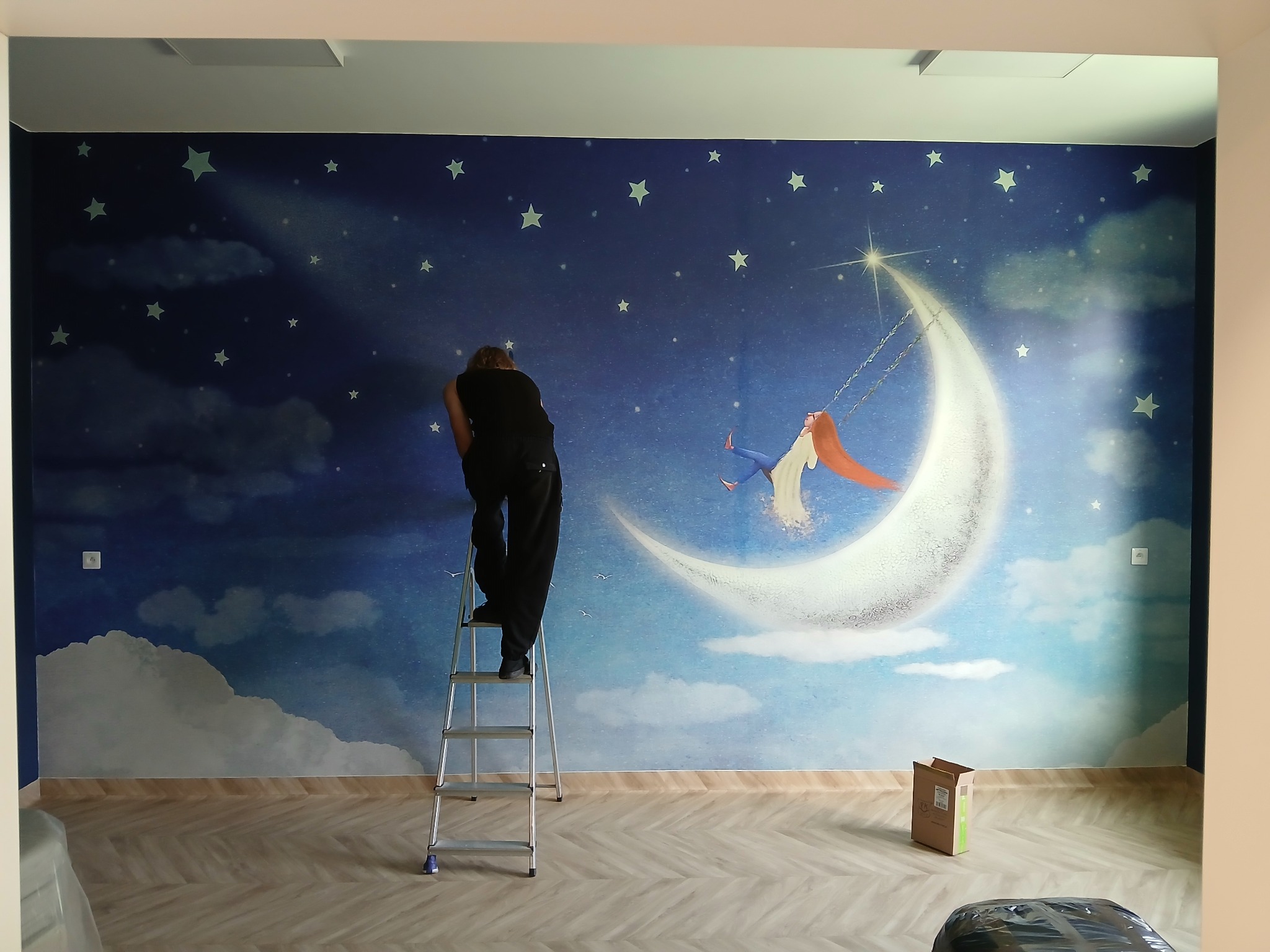 Mężczyzna na drabinie montuje fototapetę z motywem księżyca i gwiazd w pokoju. Widoczne narzędzia i karton. Jasne panele podłogowe.