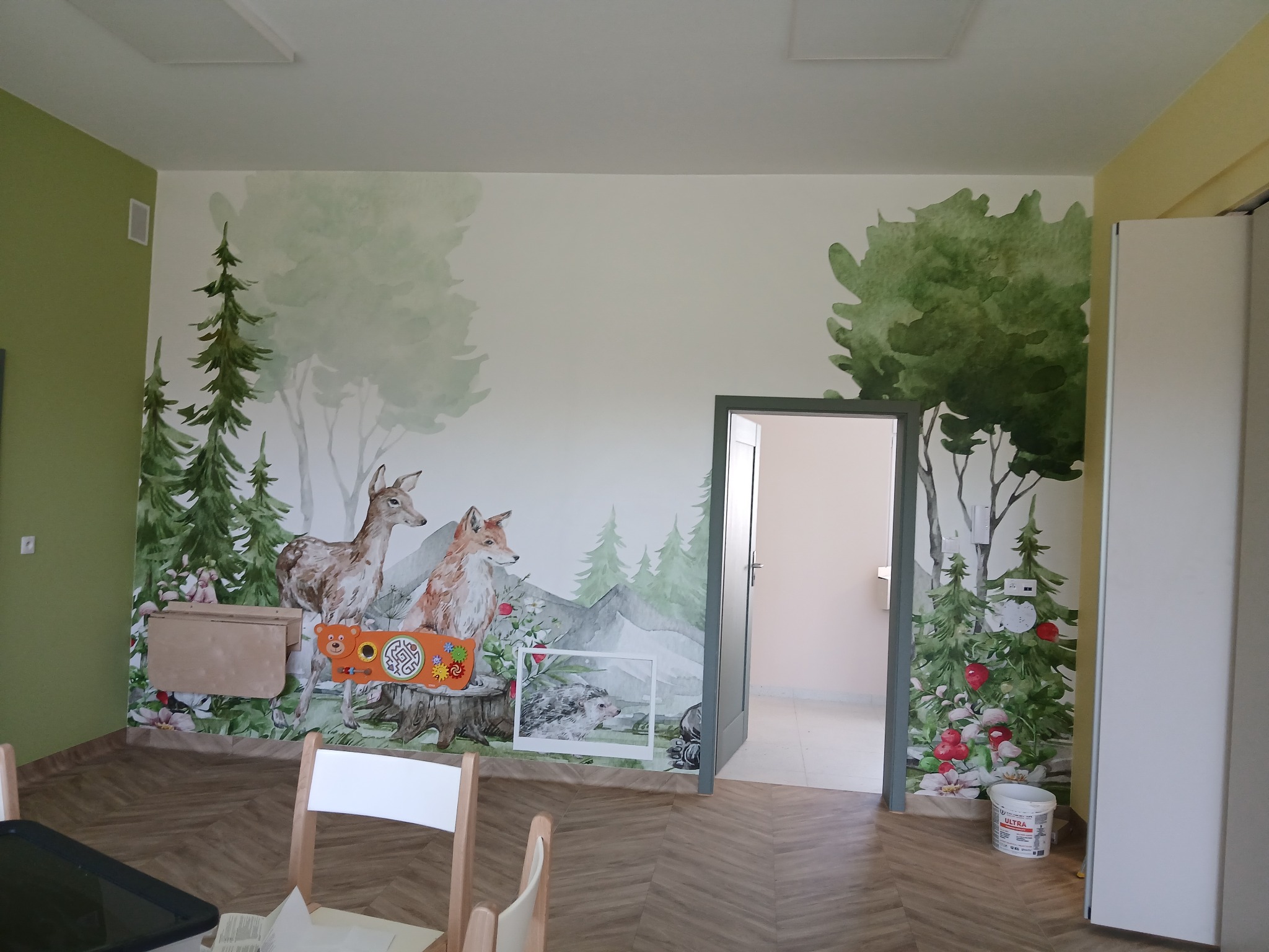 Pokój dziecięcy z malowidłami ściennymi: leśny krajobraz z jeleniem, lisem i drzewami. Na ścianie zabawki edukacyjne i obrazek z jeżem. Parkiet na podłodze.