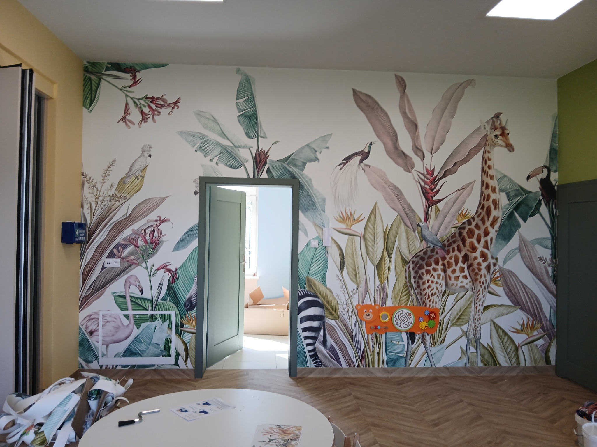 Pokój po malowaniu z motywem dżungli: żyrafa, flaming, zebra i rośliny na ścianie. Widoczne resztki tapety i stół z narzędziami. Podłoga w jodełkę.