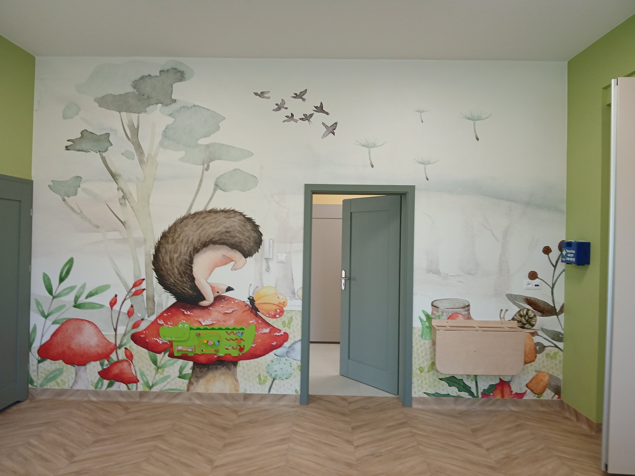Pokój dziecięcy z ręcznie malowaną ścianą: jeż na muchomorze, motyl, drzewa, ptaki, dmuchawce. Jasne kolory, drewniana podłoga, otwarte drzwi. Unikalny wzór.