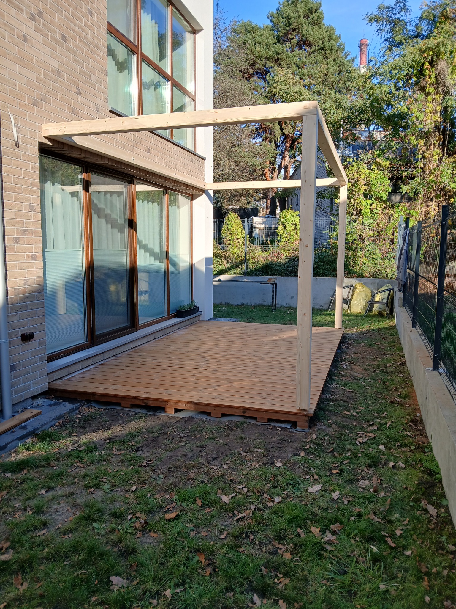 Drewniany taras przy domu w trakcie budowy, widoczne deski i konstrukcja z belek. W tle ceglana ściana budynku i zieleń ogrodu.