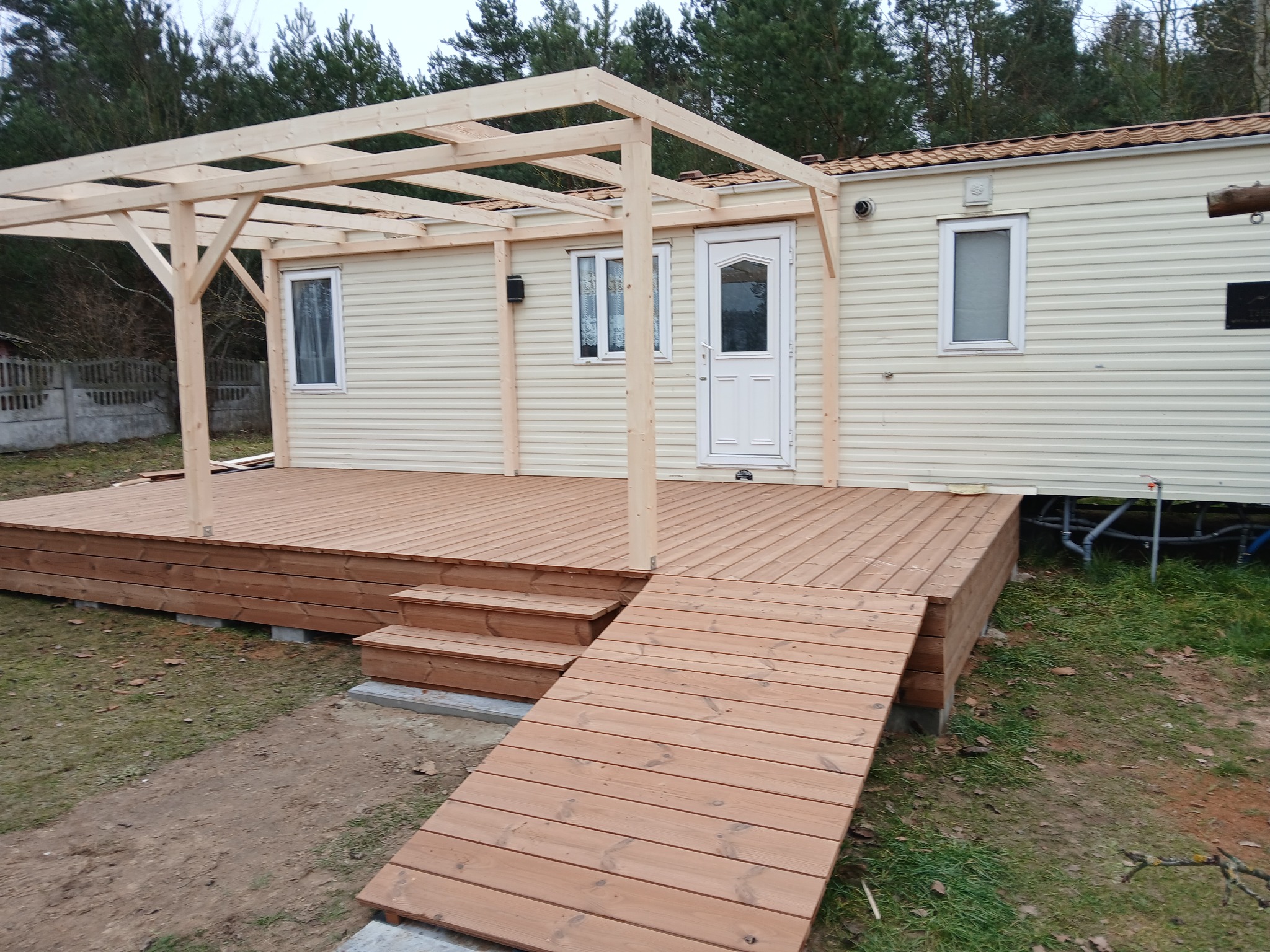 Nowy, drewniany taras z rampą i pergolą przy domu mobilnym. Jasne drewno, świeżo po budowie, widoczne elementy konstrukcyjne. Otoczenie naturalne, drzewa w tle.