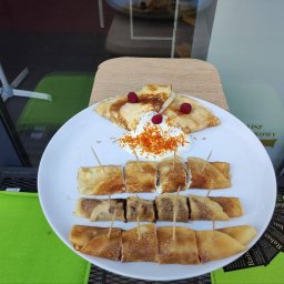 Dmitrii Baturo Best_Sonn_ - Elegancko podane naleśniki na białym talerzu z bitą śmietaną, malinami i skórką pomarańczową, pokrojone i spięte wykałaczkami, na tle szyby wystawowej.