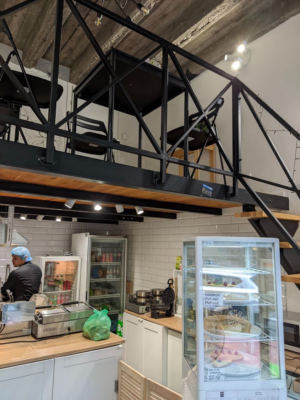 Wnętrze małej kawiarni z antresolą: widok na zaplecze z pracownikiem w czepku, chłodniczą witrynę z ciastami, ekspresy do kanapek i gofrownicę na blacie.