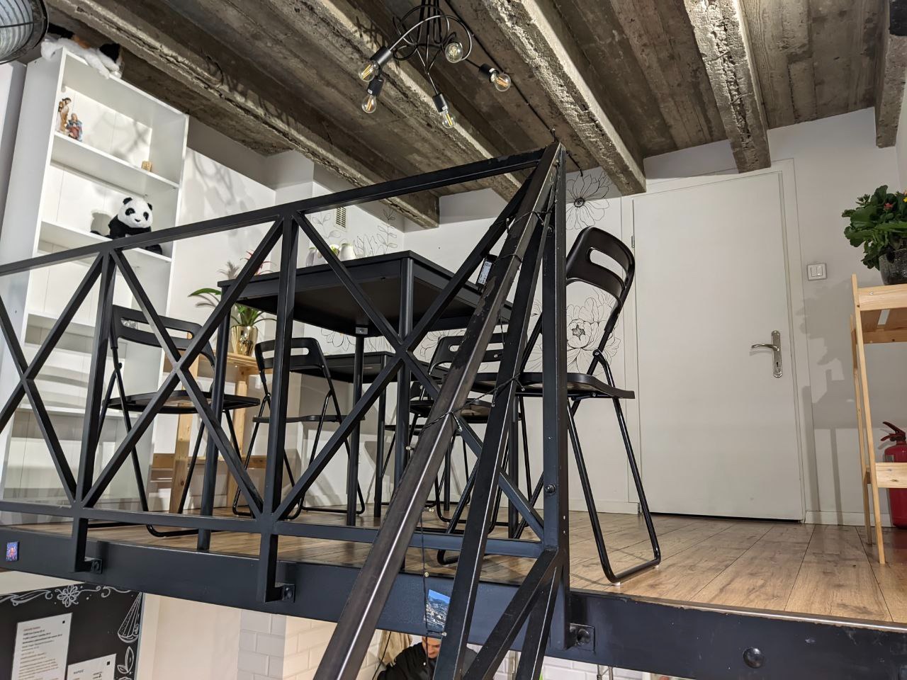 Nowoczesna przestrzeń biurowa z antresolą, czarnym metalowym balustradą i stołem z krzesłami, widok na białe ściany z dekoracyjnymi naklejkami i drewnianą podłogę.