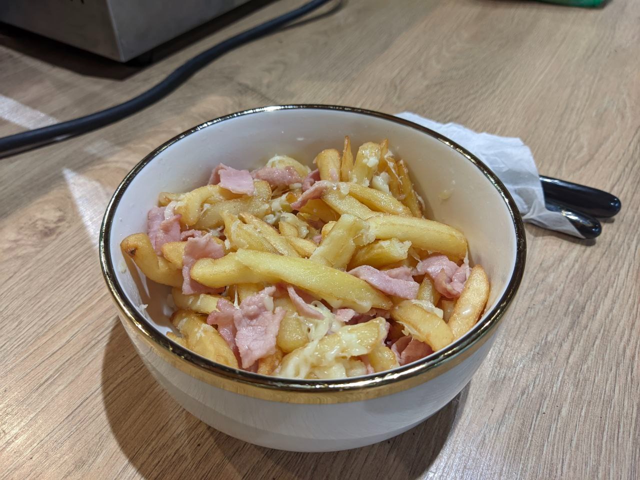 Frytki z szynką i serem w białej miseczce ze złotym rantem, widok z góry, obok leżą sztućce.