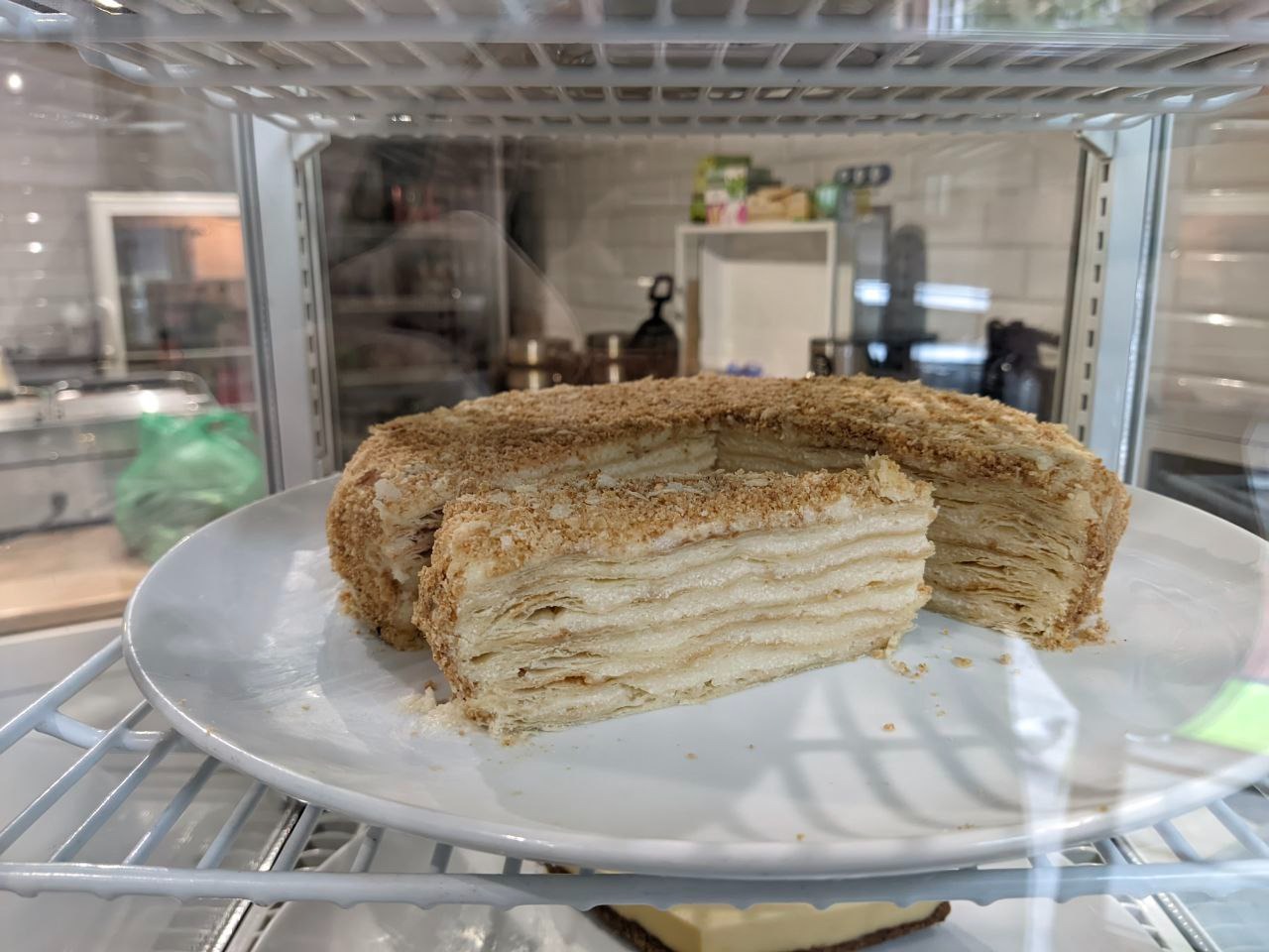 Tort Napoleonka z widocznymi warstwami ciasta francuskiego i posypką z kruszonki, na białym talerzu w witrynie chłodniczej.