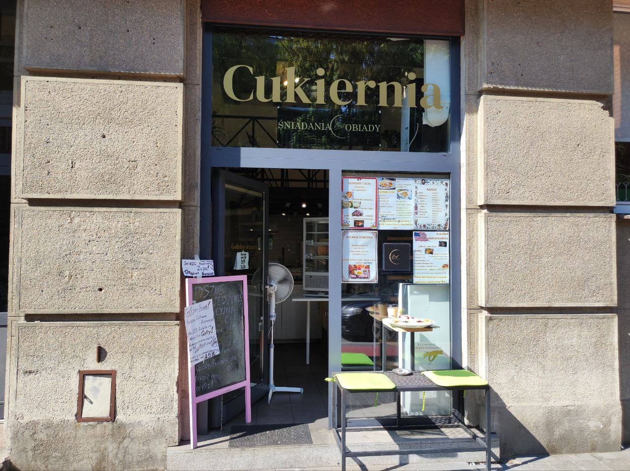 Wejście do cukierni z szyldem 'Cukiernia Śniadania Obiady', menu na szybie, stolik z dwoma krzesłami i tablicą z ofertą dnia.