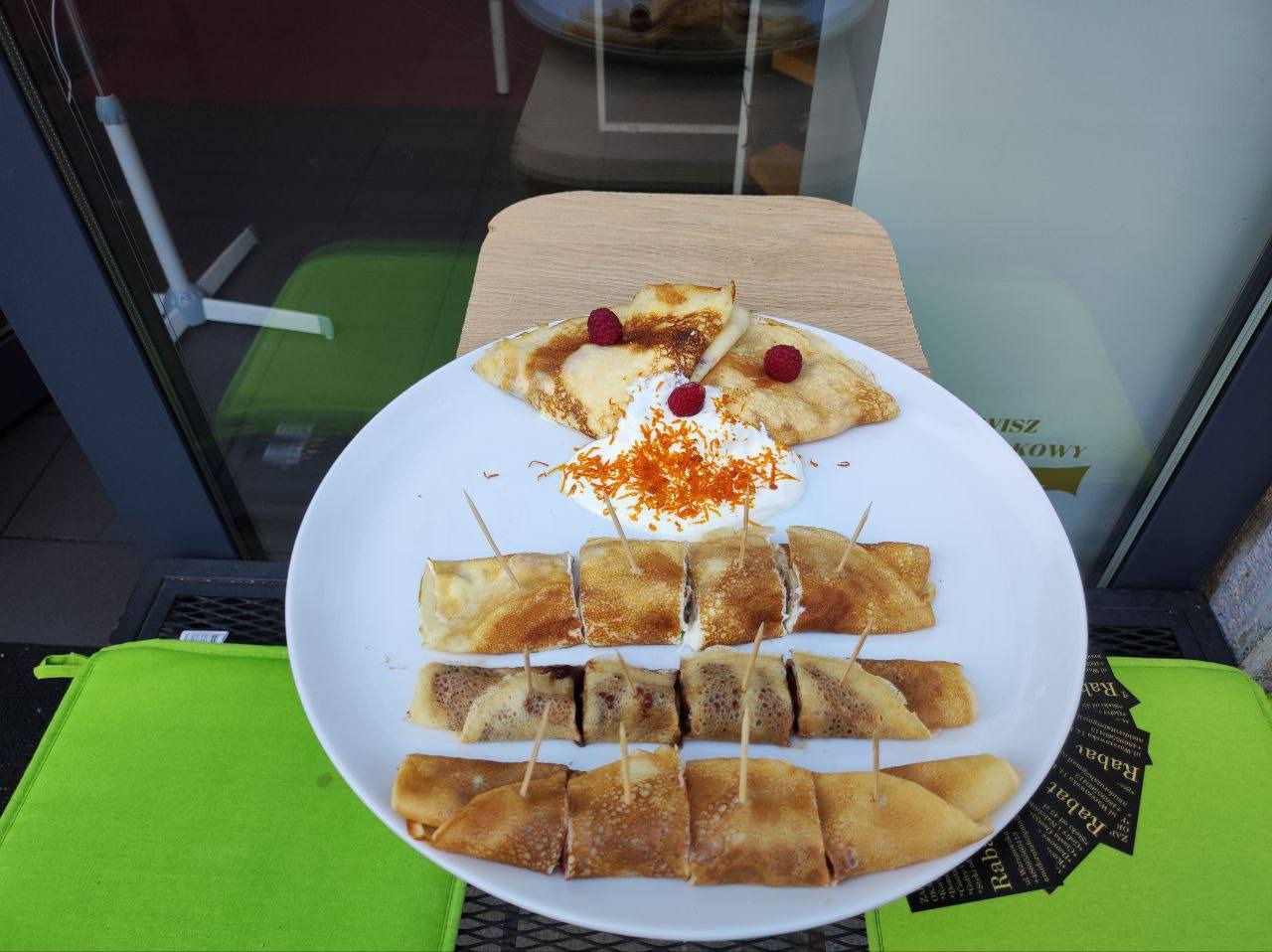 Elegancko podane naleśniki na białym talerzu z bitą śmietaną, malinami i skórką pomarańczową, pokrojone i spięte wykałaczkami, na tle szyby wystawowej.