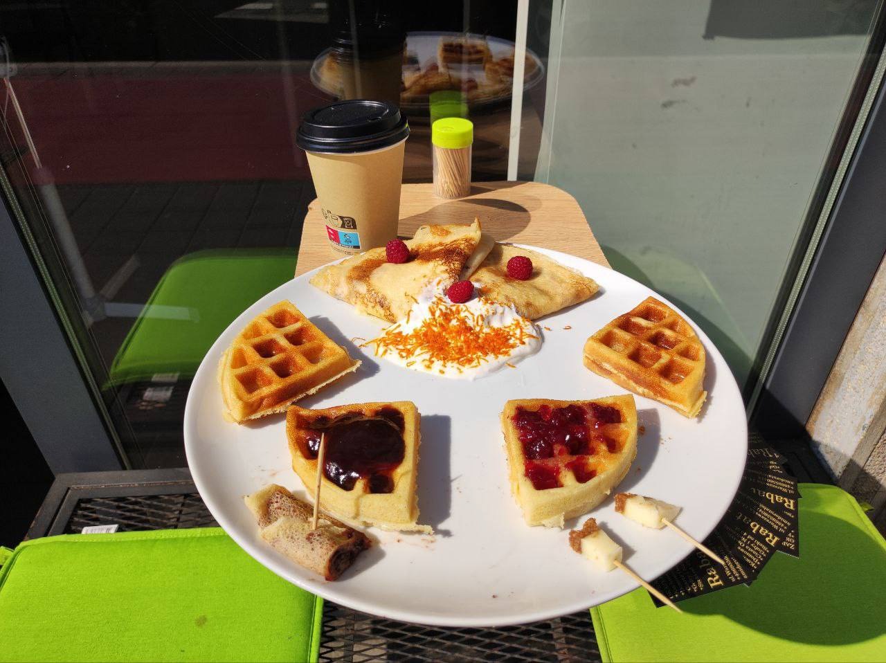 Talerz z naleśnikami z bitą śmietaną i malinami, goframi, kawałkami ciasta z dżemem, roladką i kawą na wynos, ustawiony na stoliku z zielonymi krzesłami w tle.