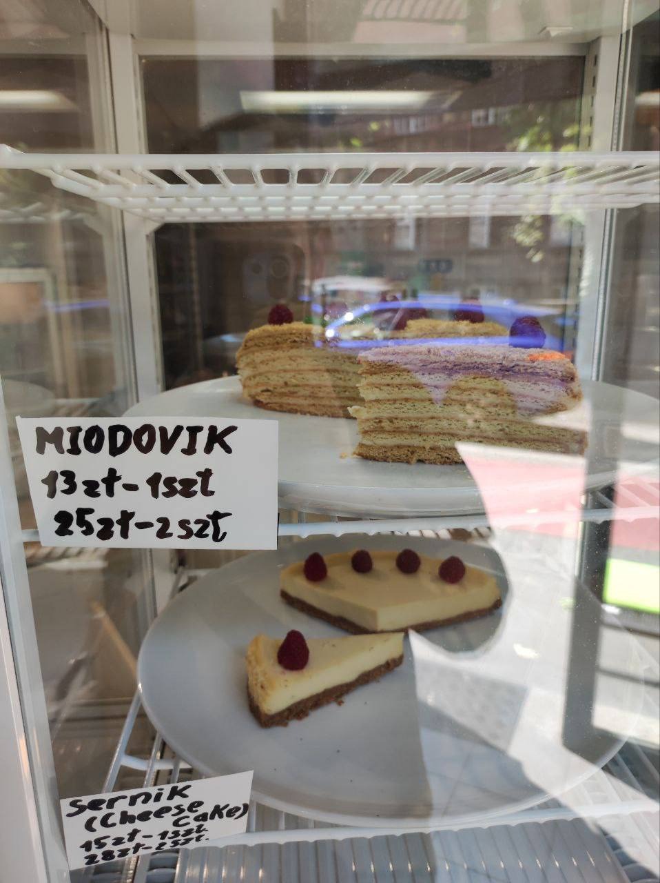 Dwa rodzaje ciast w chłodniczej witrynie: Miodownik z maliną na wierzchu oraz sernik nowojorski z malinami, obok ceny wypisane na kartkach.