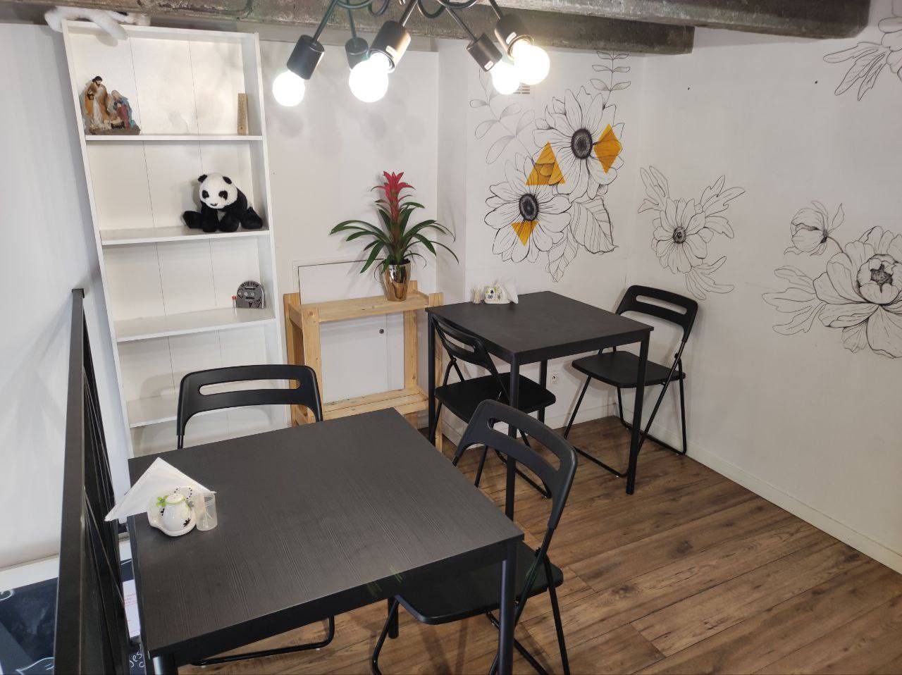 Wnętrze kawiarni z dwoma czarnymi stolikami i krzesłami, na ścianie rysunki kwiatów, na półce panda i roślina w doniczce.