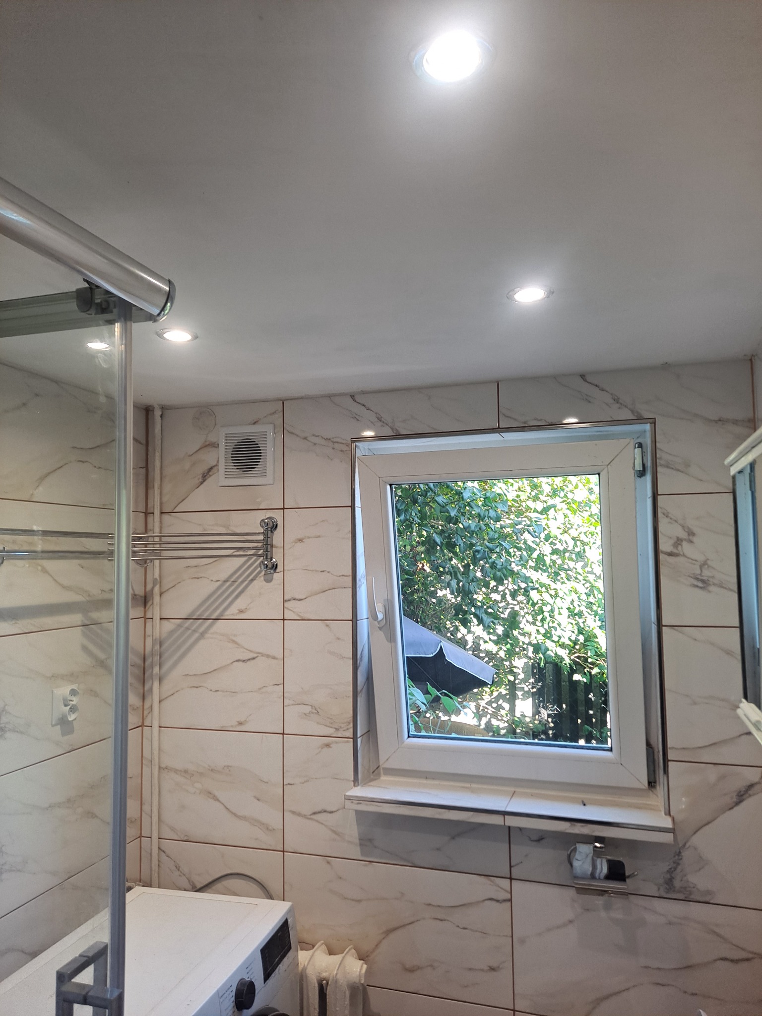 Wykończona łazienka z kabiną prysznicową, oknem wychodzącym na zieleń i marmurkowymi płytkami na ścianach, oświetlona punktowymi lampami sufitowymi, z pralką i grzejnikiem widocznymi w dolnej...