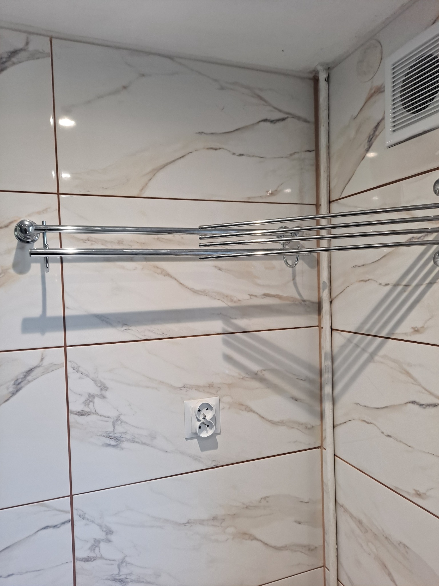 Ściana łazienki wyłożona białymi płytkami imitującymi marmur, z zamontowanym chromowanym wieszakiem na ręczniki, widoczny gniazdko elektryczne i kratka wentylacyjna.