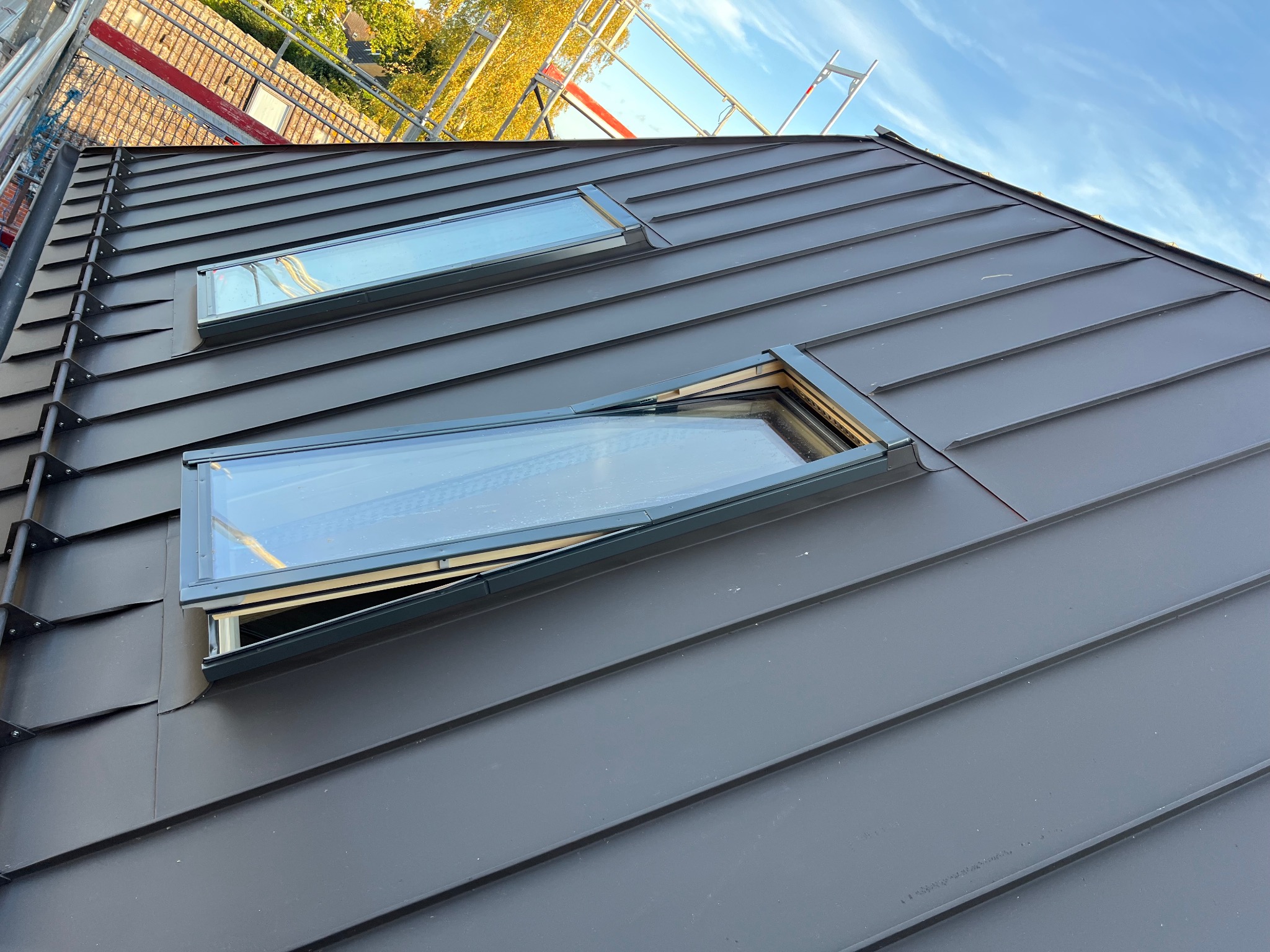 Grafitowy dach z blachy na rąbek stojący z dwoma oknami dachowymi, jedno uchylone, widok z dołu, na tle rusztowania i błękitnego nieba.