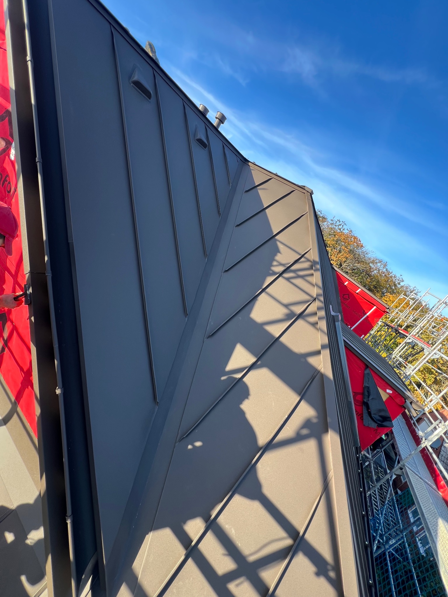 Szary dach z blachy na rąbek stojący marki Prefa, widok z góry, częściowo widoczne rusztowanie i błękitne niebo.