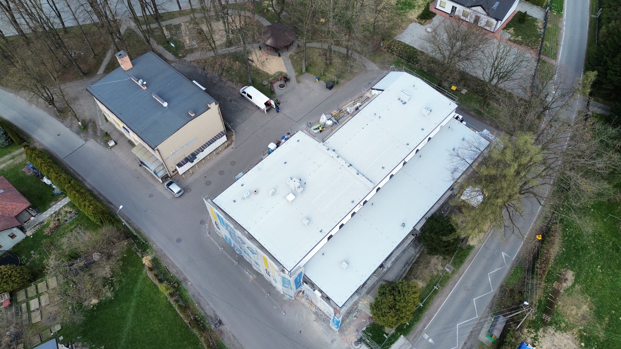 Widok z drona na budynek z białym dachem, gdzie trwają prace dekarskie, widoczne rusztowania i materiały budowlane, otoczenie to tereny zielone i zabudowa miejska.