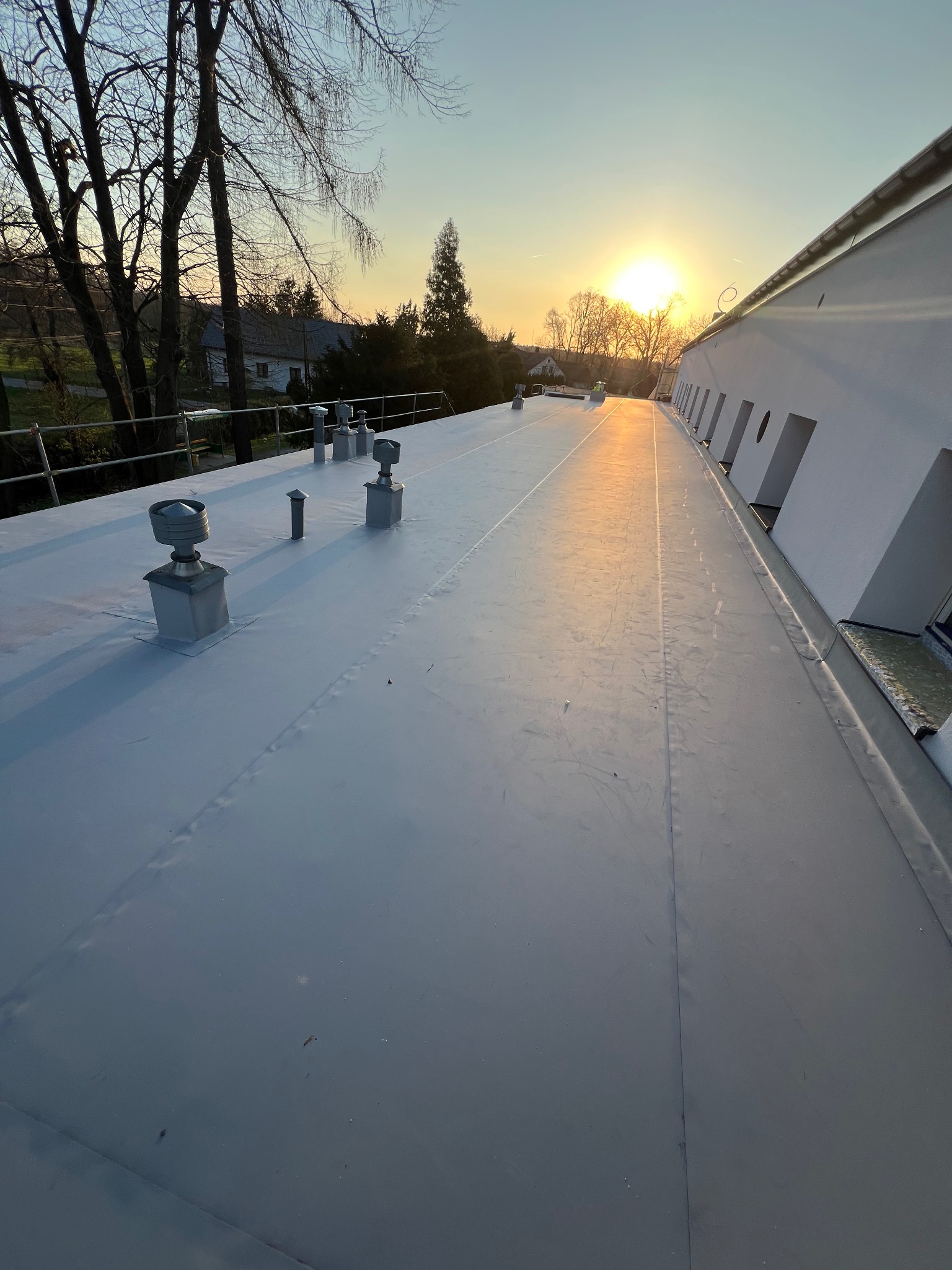 Szary dach pokryty membraną TPO BAUDER, z kominkami wentylacyjnymi, oświetlony promieniami zachodzącego słońca.