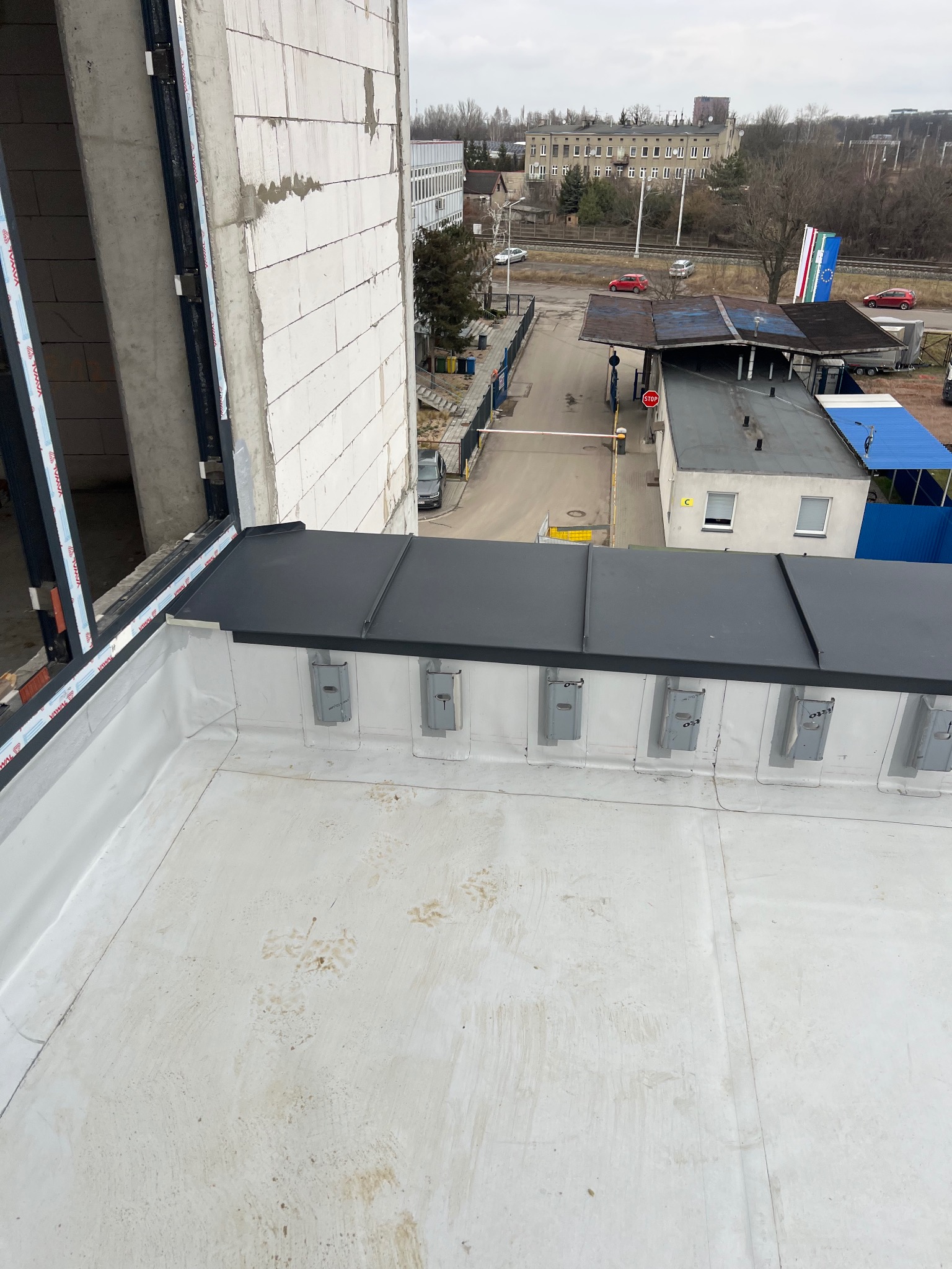 Widok z góry na płaski dach z elementami obróbki blacharskiej i membraną dachową, z widocznym fragmentem budynku w stanie surowym otwartym i zabudowaniami w tle.