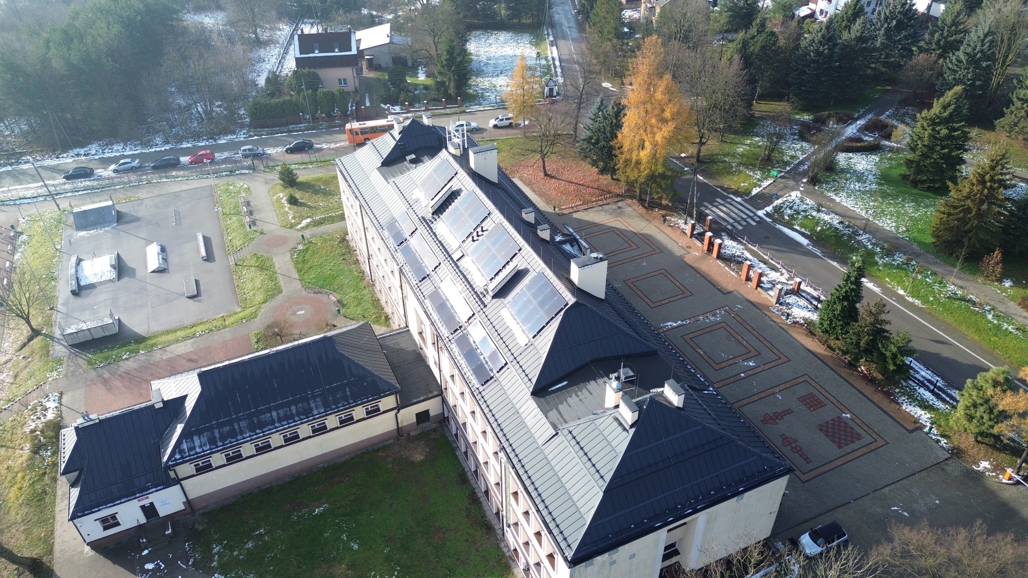 Widok z góry na budynek z ciemnym dachem pokrytym panelami słonecznymi, obok skatepark i drzewa z resztkami śniegu.
