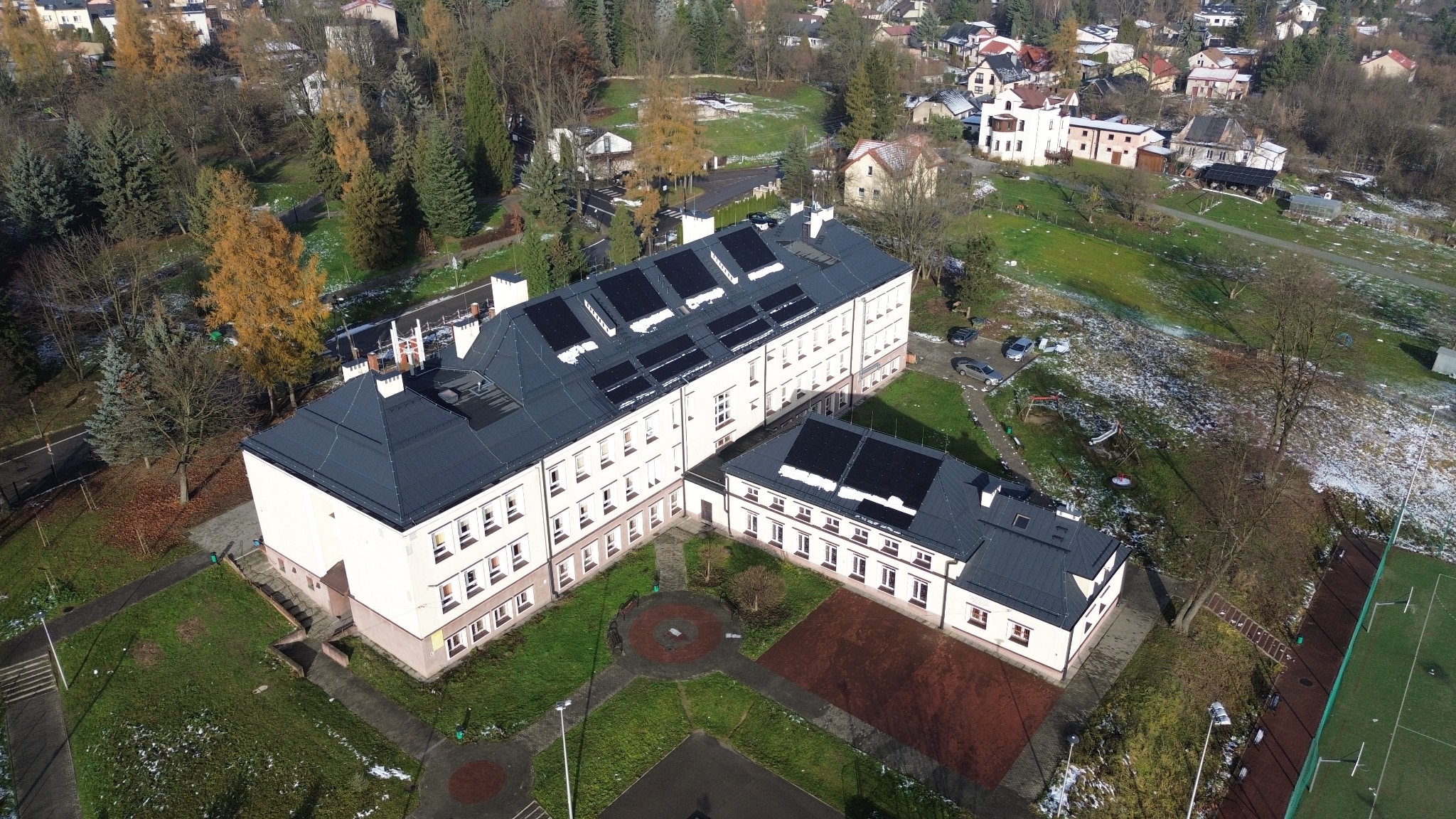Widok z lotu ptaka na duży budynek z ciemnym dachem, na którym zamontowano panele słoneczne; w tle widoczne zabudowania mieszkalne i drzewa.