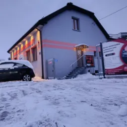 Zimowy widok na budynek warsztatu samochodowego z napisem 'Warsztat Samochodowy' i logo 'Profi Auto', z zaparkowanym autem częściowo zasypanym śniegiem oraz tablicą reklamową usług.