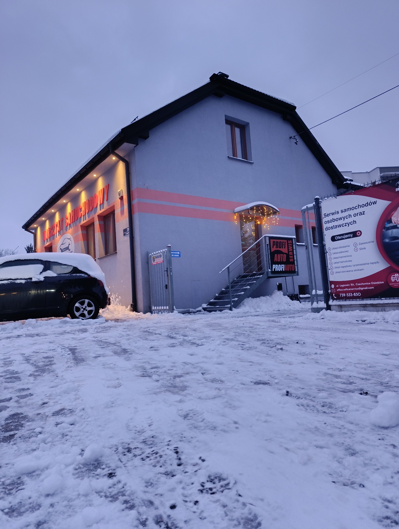 Zimowy widok na budynek warsztatu samochodowego z napisem 'Warsztat Samochodowy' i logo 'Profi Auto', z zaparkowanym autem częściowo zasypanym śniegiem oraz tablicą reklamową usług.