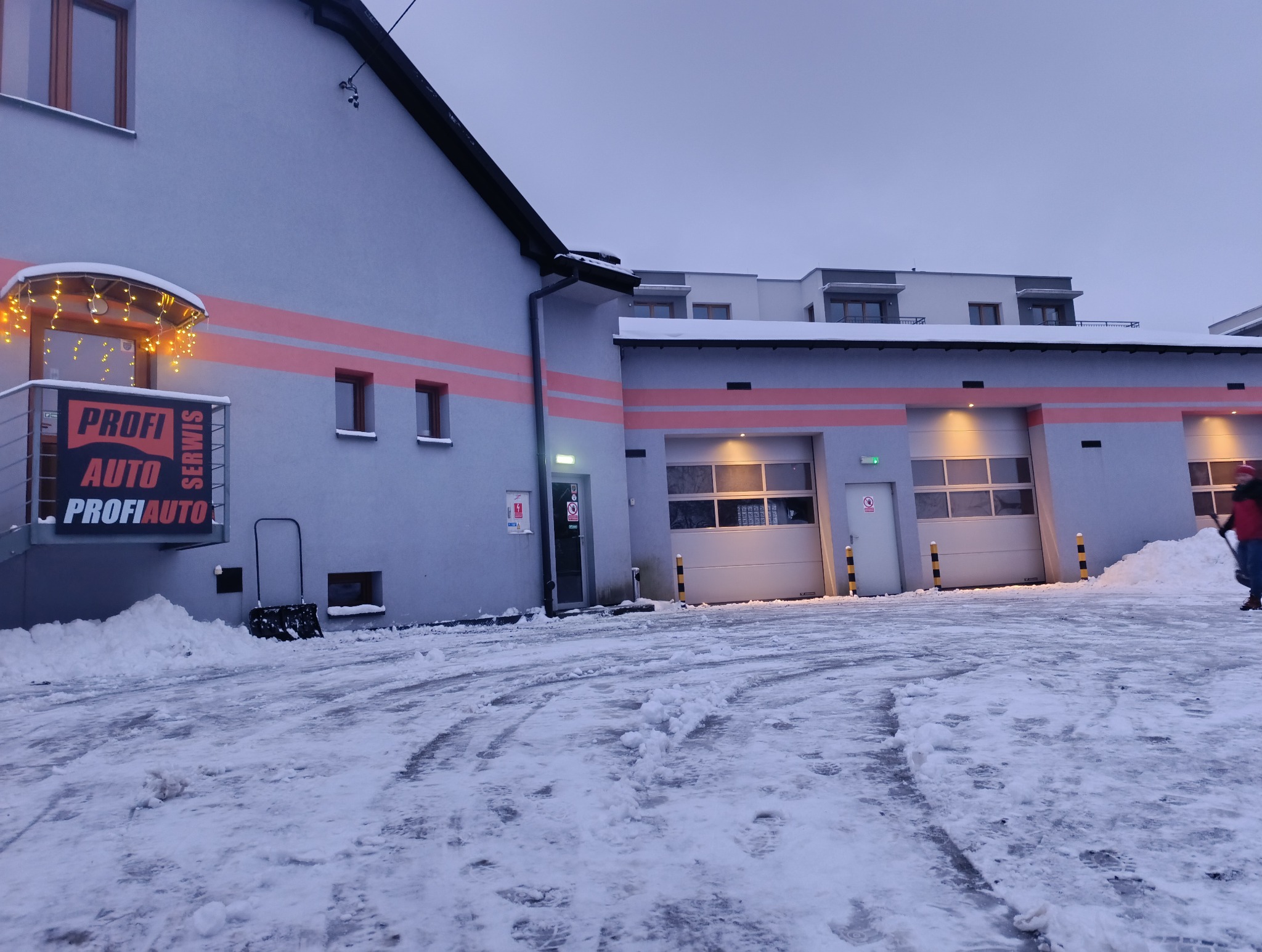 Zimowy widok na budynek warsztatu samochodowego z logo 'PROFI AUTO SERWIS', oświetlonym wejściem i odśnieżonym placem przed bramami garażowymi.