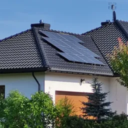 Dom jednorodzinny z panelami fotowoltaicznymi na dachu z dachówki ceramicznej, antena telewizyjna na kominie, widok z poziomu trawnika.