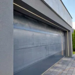 Nowoczesna, szara brama garażowa segmentowa z oświetleniem LED wzdłuż górnej krawędzi i po bokach, widoczny fragment kostki brukowej i przewód elektryczny na ziemi.