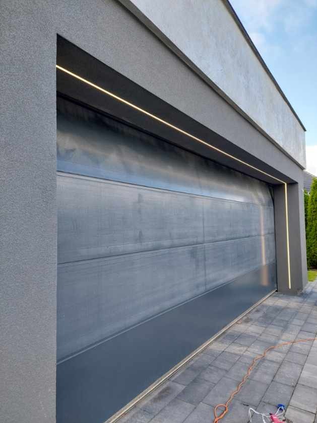 Nowoczesna, szara brama garażowa segmentowa z oświetleniem LED wzdłuż górnej krawędzi i po bokach, widoczny fragment kostki brukowej i przewód elektryczny na ziemi.