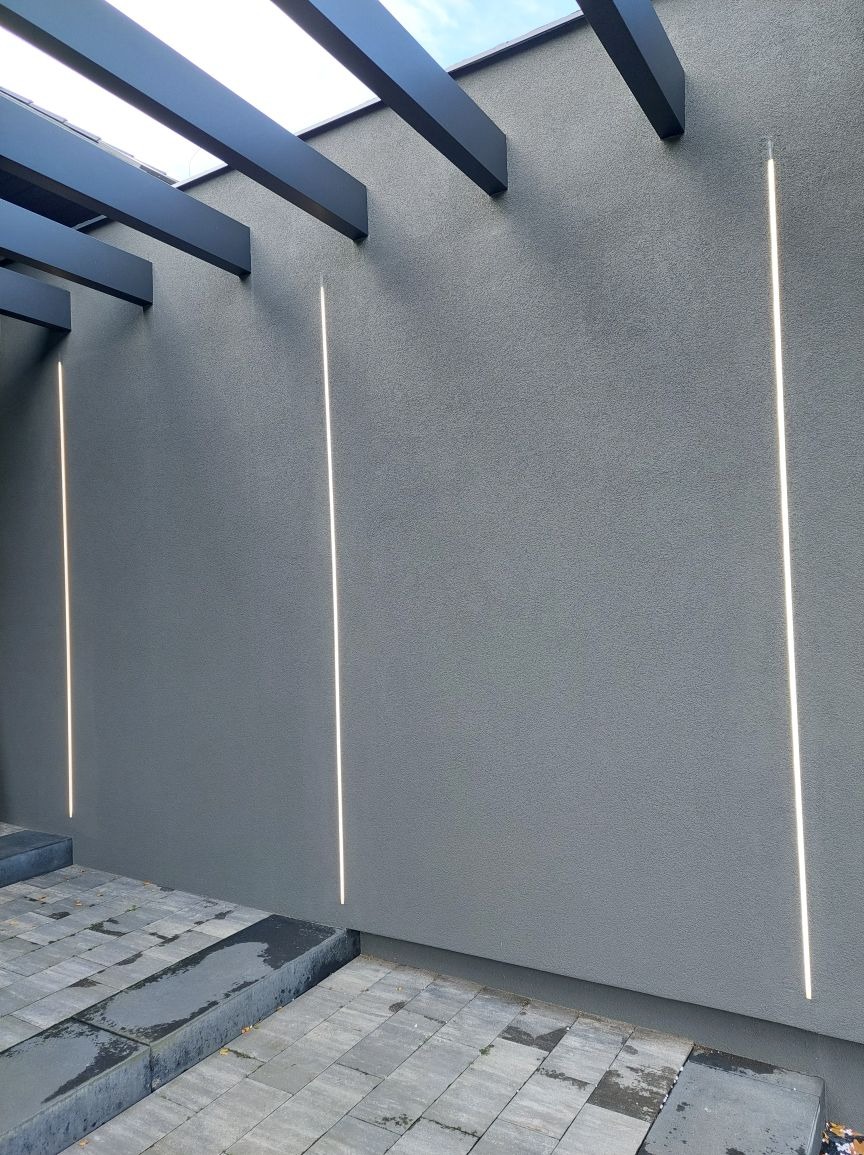 Elewacja budynku z szarego tynku z wkomponowanymi pionowymi liniami świetlnymi LED, widoczne fragmenty czarnej konstrukcji zadaszenia i szara kostka brukowa na dole.