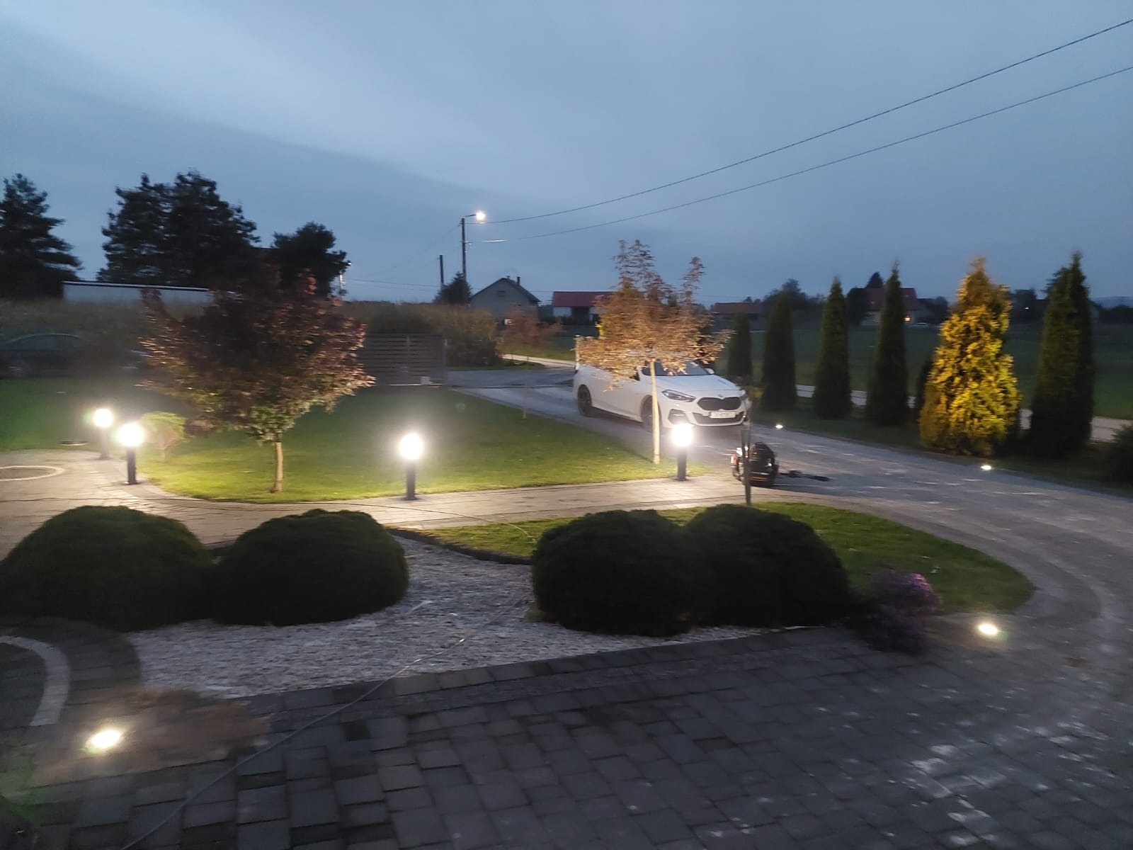 Oświetlony podjazd z lampami ogrodowymi wzdłuż ścieżki i trawnika, biały samochód w tle, wieczorny krajobraz.