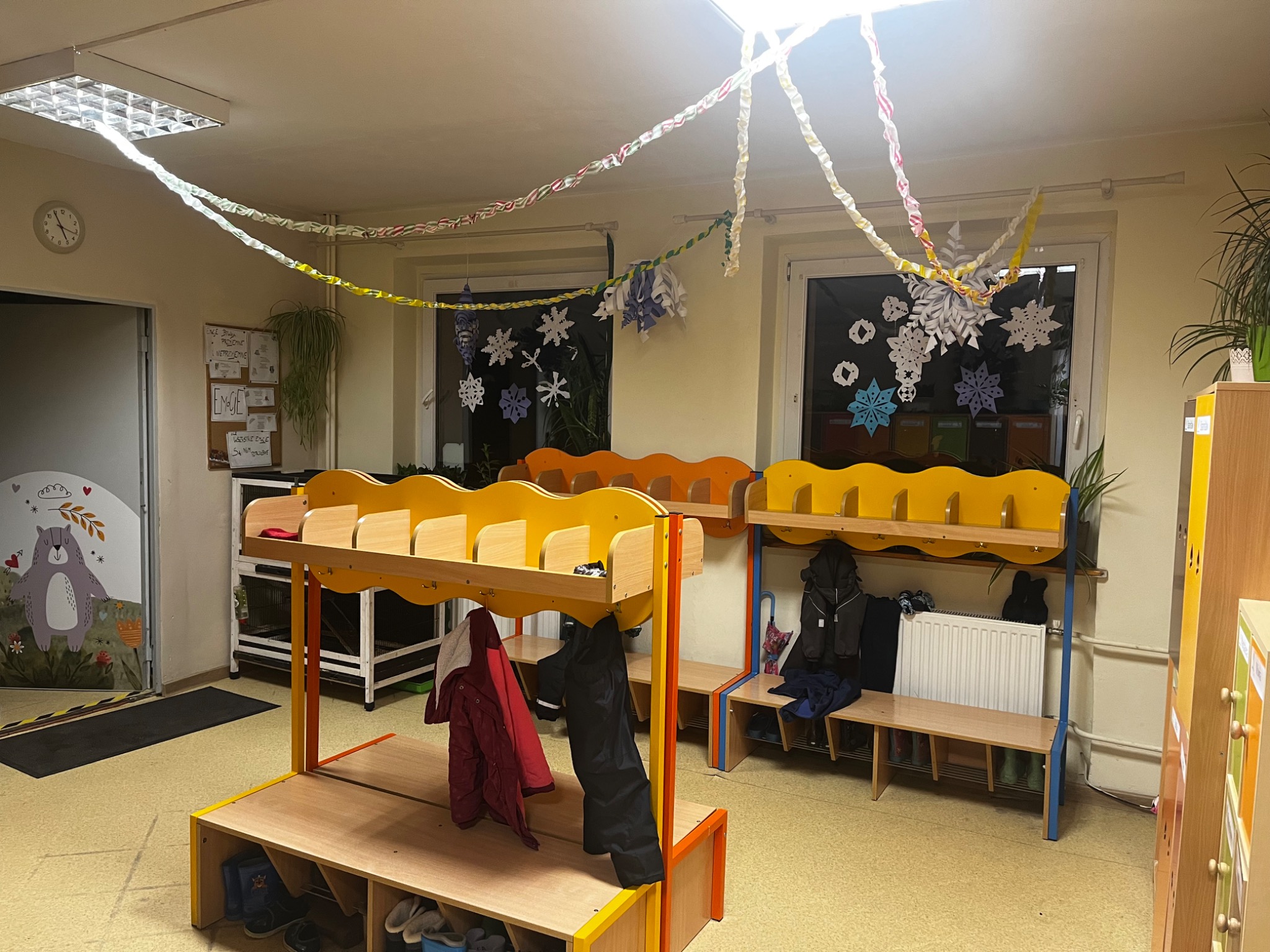 Wnętrze szatni przedszkolnej z żółto-pomarańczowymi szafkami na ubrania, udekorowane papierowymi łańcuchami i śnieżynkami na oknach.