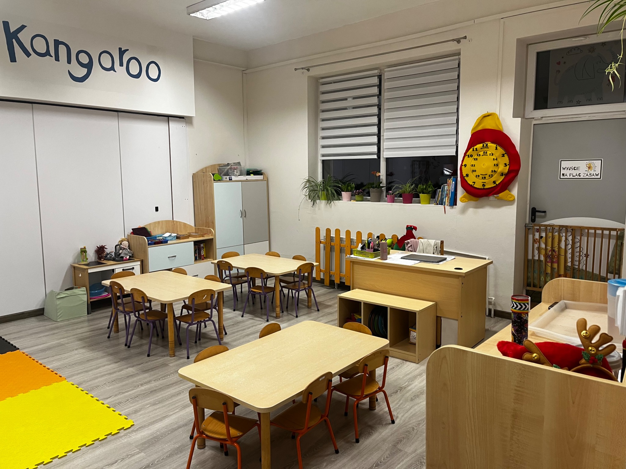 Jasne wnętrze sali przedszkolnej z drewnianymi stolikami i krzesłami, kolorową podłogą z puzzli, dekoracyjnym zegarem w kształcie czerwonego ptaka na ścianie i napisem 'Kangaroo' na górze ściany.