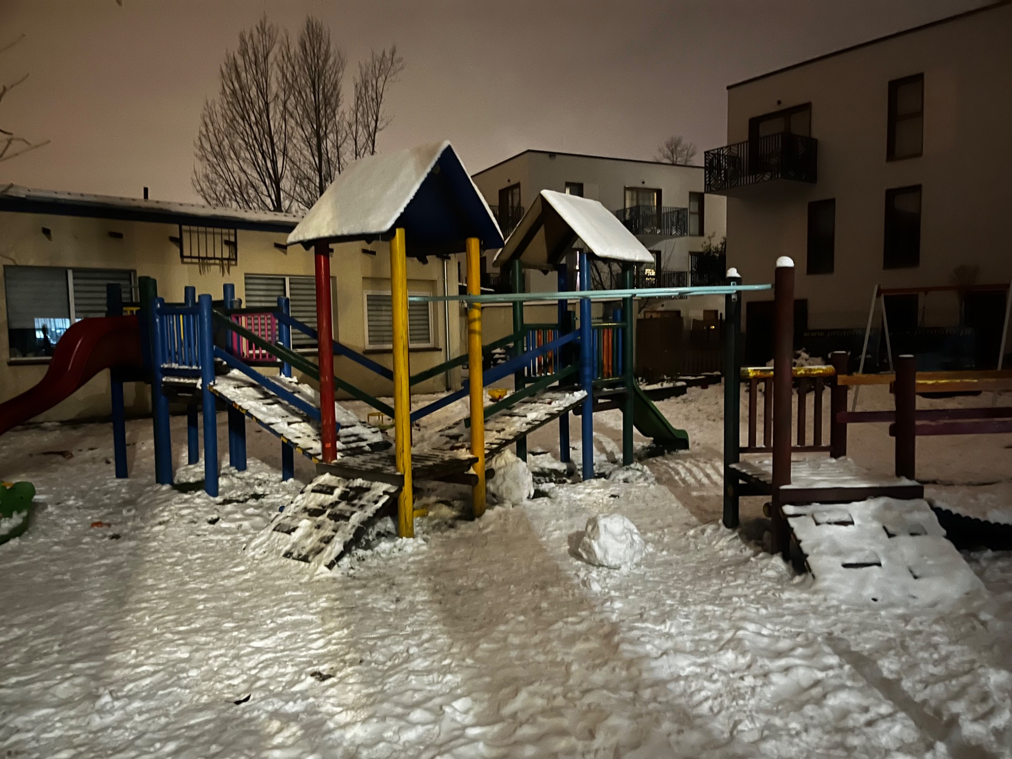 Zimowy plac zabaw z drewnianą konstrukcją pokrytą śniegiem, w tle budynki mieszkalne oświetlone nocą.