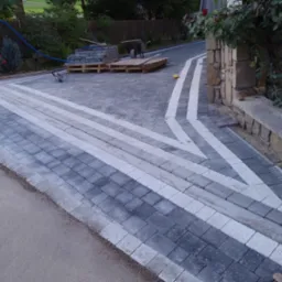 Ułożona nawierzchnia z kostki brukowej w odcieniach szarości z białymi pasami, prowadząca do kamiennego murku z rosnącymi różami. Na paletach w tle widoczne zapasy kostki i taczka.