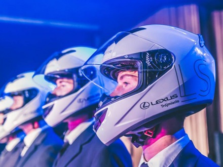 Rząd mężczyzn w garniturach i białych kaskach motocyklowych z logo Lexus Trójmiasto podczas prezentacji produktu na evencie firmowym.