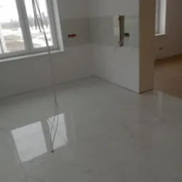 Jasne, błyszczące płytki podłogowe imitujące marmur w surowym wnętrzu z częściowo położonymi płytkami ściennymi i wystającymi kablami elektrycznymi.