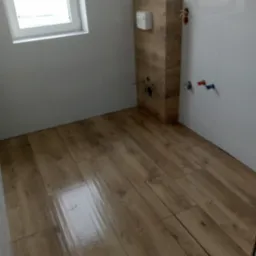 Wykończona łazienka z drewnopodobną podłogą, zabudową stelaża WC i białymi płytkami na ścianach, widoczne instalacje wodne.